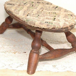 May include: An antique wooden footstool with a worn, oval-shaped top. The top is covered in a faded, patterned fabric with beige, green, and black hues. The stool has four turned legs, and the wood has a warm, aged appearance.
