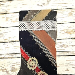 May include: A holiday stocking constructed from various fabric patches in black, red, brown, and houndstooth patterns. The pieces are joined by visible white decorative stitching. A wide strip of white lace and two circular lace doilies are attached.