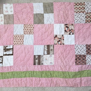 May include: A rectangular patchwork quilt featuring a variety of fabric squares in shades of pink, brown, white, and green. The design incorporates floral, paisley, striped, and polka dot patterns, finished with a solid green horizontal border.