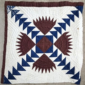 May include: A square quilt block featuring a central brown diamond surrounded by four brown fan-shaped motifs. The design includes blue triangles arranged in geometric patterns against a white background with intricate quilted stitching.