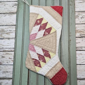 May include: A quilted Christmas stocking featuring a geometric patchwork design in shades of cream, tan, pale yellow, and deep red. The stocking has a white hanging loop at the top and is constructed with visible stitching throughout the fabric panels.