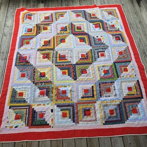 May include: A rectangular quilt featuring a log cabin pattern composed of numerous square blocks. Each block contains a central red square surrounded by concentric strips of various patterned and solid fabrics in blue, yellow, green, and white, finished with a wide red border.