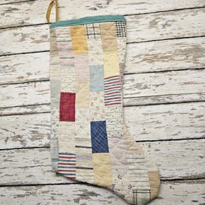 May include: A patchwork Christmas stocking with a variety of fabric patterns and colors, including red, blue, yellow, and white. The stocking has a teal cuff and a gold hanging loop. The design is quilted.