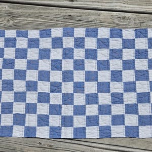 May include: A rectangular quilted textile featuring a repeating checkerboard pattern of alternating solid blue and white squares. The fabric has a visible stitched texture throughout, creating a soft, slightly puckered appearance across the surface.