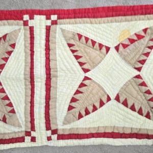 May include: A rectangular quilted textile featuring a geometric pattern in cream, tan, and red. The design includes triangular shapes with serrated red edges arranged in star-like formations, bordered by red and cream bands with small checkered accents.