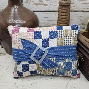 May include: A square patchwork pincushion featuring a grid of small fabric squares in shades of blue, white, tan, and red. A blue fabric strip is wrapped horizontally around the center, secured with a clear rectangular plastic buckle.