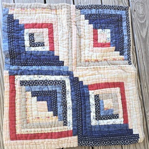 May include: A square fabric quilt featuring a log cabin pattern composed of four distinct blocks. The design incorporates various textile strips in shades of navy blue, red, cream, and white, with assorted floral, plaid, and polka dot prints.