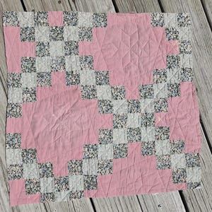 May include: A square quilt featuring a repeating pattern of solid dusty pink fabric blocks and smaller squares of floral print fabric in shades of blue, pink, and white. The quilt surface displays intricate stitched star motifs.