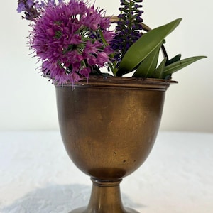 Könnte beinhalten: Eine bronzefarbene, kelchförmige Vase enthält einen kleinen Strauß aus lila Blumen und grünen Blättern. Die Vase hat eine abgerundete Schale, einen Sockel und einen leicht ausgestellten Rand. Die Blumen bilden einen Kontrast zum metallischen Finish der Vase.