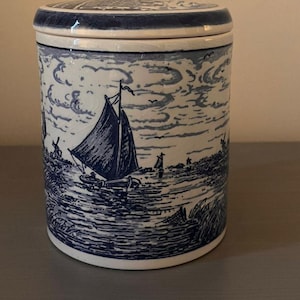 May include: A blue and white ceramic storage jar with a lid, depicting a nautical scene with sailboats and a coastal landscape. The jar's design is reminiscent of traditional Delftware, suitable for kitchen or home decor.