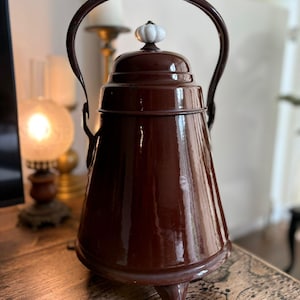 Puede incluir: Una tetera vintage de metal marrón oscuro con un acabado brillante. Cuenta con un asa curva, un pomo blanco y marrón en la tapa y descansa sobre tres patas pequeñas. La tetera tiene un diseño clásico y antiguo.