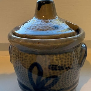 May include: A small, lidded ceramic jar with a blue and brown glaze. The jar has a textured surface and a blue floral design. The lid has a small handle. The jar has two small handles on the sides.