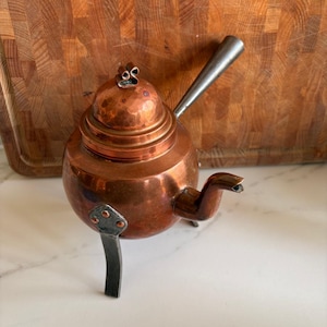 Könnte beinhalten: Eine kleine Kupferteekanne mit gehämmerter Oberfläche und einem schwarzen Metallgriff und -beinen. Die Teekanne hat einen runden Korpus und einen gewölbten Deckel.