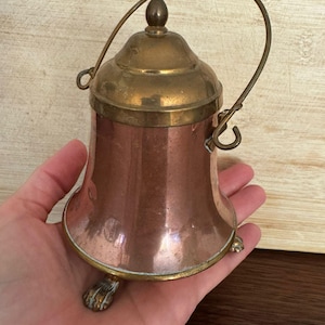 Puede incluir: Un recipiente de cobre y latón con tapa y asa. El recipiente tiene forma de campana con un borde de latón y un pie pequeño. El asa es de latón y se arquea hacia arriba. El recipiente es probablemente vintage.