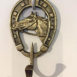 Peut inclure: Un crochet mural en métal de couleur bronze en forme de fer à cheval avec une tête de cheval au centre. Le crochet est fixé à une pièce en plastique grise pour l'accrochage. Le crochet est conçu pour suspendre des objets.