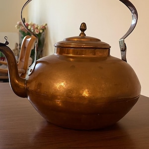 Puede incluir: Una tetera de cobre con cuerpo redondeado, asa curva y tapa con pequeño pomo. La tetera tiene un pico y probablemente se utiliza para hervir agua. El cobre tiene un color marrón rojizo cálido. Diseño clásico.