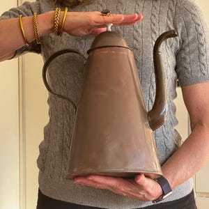 Puede incluir: Una cafetera de cobre grande con un pico largo y un asa. La cafetera tiene un acabado deslustrado y está sostenida por las manos de una persona.