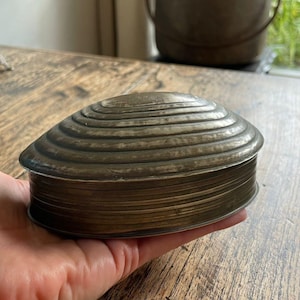 Pode incluir: Uma caixa antiga em forma de concha, feita de metal envelhecido. A tampa apresenta um design canelado e a caixa tem uma aparência manchada. A caixa é segurada em uma mão, mostrando seu tamanho e detalhes.