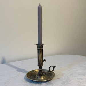 Puede incluir: Un candelabro de latón antiguo con una vela gris. El candelabro tiene una base redonda, un tallo central y un pequeño asa. La vela es de color gris claro y se inserta en la parte superior del candelabro. El candelabro está sobre un mantel blanco.