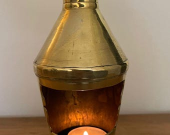 Puede incluir: Un portavelas de latón con una vela de té encendida en su interior. El portavelas tiene forma cónica con una parte superior acampanada y una pequeña abertura para que brille la luz. El portavelas está sobre una superficie de madera.