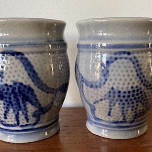 Puede incluir: Dos tazas pequeñas de cerámica blanca con diseños azules y blancos. Las tazas tienen una forma redondeada y un labio ligeramente acampanado.