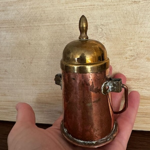 Puede incluir: Un pequeño recipiente antiguo de cobre y latón con tapa y asa. El recipiente tiene un cuerpo cilíndrico con una base redondeada y una tapa abovedada. El asa es un pequeño lazo rectangular. El recipiente se sostiene en una mano.