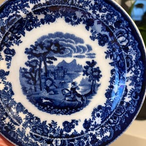 May include: A round, white and blue decorative plate. The plate features a central landscape scene with a person on horseback, trees, and a building. The border has a floral design. The plate is made of ceramic.