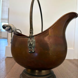 Op de afbeelding: Een koperkleurige metalen kolenkit met een gebogen tuit en een handvat. De kolenkit heeft een ronde basis en is versierd met een leeuwenkop op het bevestigingspunt van het handvat. Het handvat is gebogen en van hetzelfde metaal.