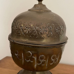 Könnte beinhalten: Eine Vintage-Schale mit Deckel in Bronzefarbe und floralen Mustern. Die Schale hat einen abgerundeten Boden und eine dekorative Spitze auf dem Deckel. Das Äußere weist geprägte Blumendesigns auf, die zu ihrer antiken Ästhetik beitragen.