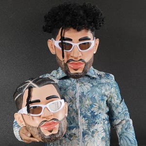 Op de afbeelding: Een persoon draagt een jas met een blauw bloemenpatroon en houdt een wit plastic masker vast van een persoon met een baard en zwart haar. Het masker heeft een witte zonnebril en een zwarte baard.