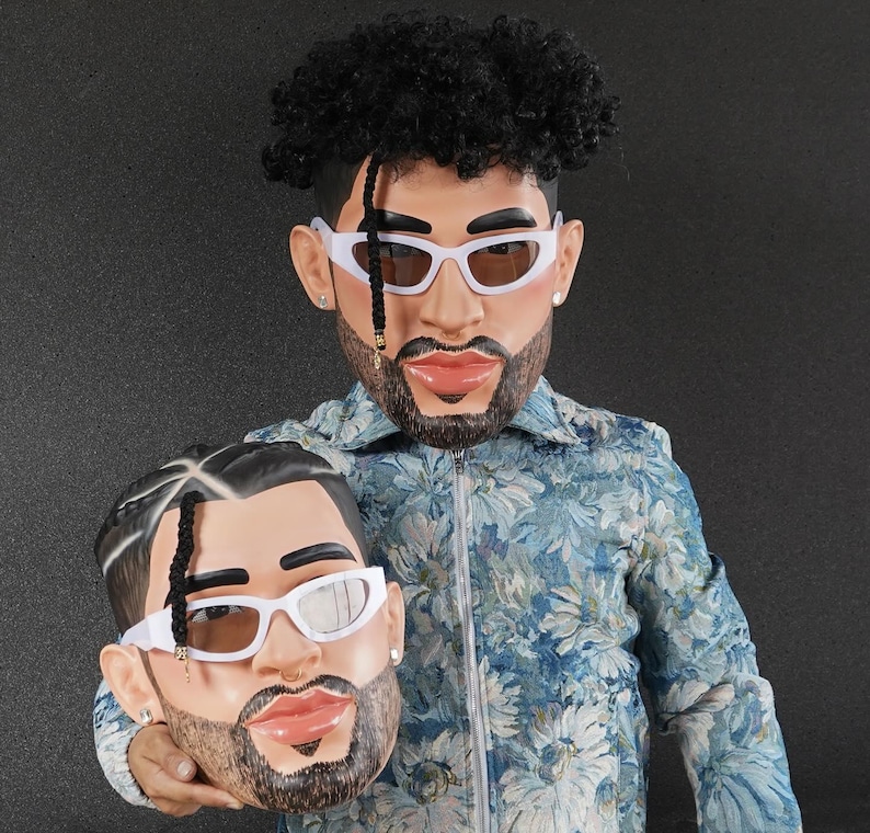 Op de afbeelding: Een persoon draagt een jas met een blauw bloemenpatroon en houdt een wit plastic masker vast van een persoon met een baard en zwart haar. Het masker heeft een witte zonnebril en een zwarte baard.
