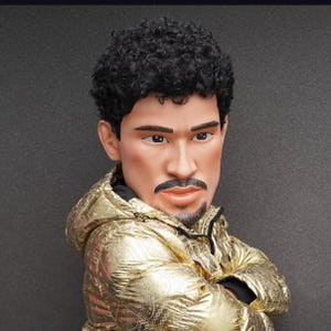 Op de afbeelding: Een man met kort, krullend zwart haar draagt een gouden jas met een rits. Hij kijkt naar de zijkant met zijn armen gekruist.