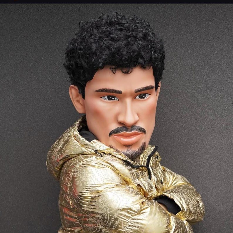 Op de afbeelding: Een man met kort, krullend zwart haar draagt een gouden jas met een rits. Hij kijkt naar de zijkant met zijn armen gekruist.