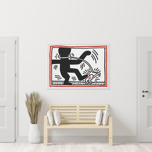 May include: A framed artwork featuring a black and white graphic of a figure with a leash, walking another figure. The artwork has a red border and is displayed above a wooden bench with white pillows. The room has white doors and walls.