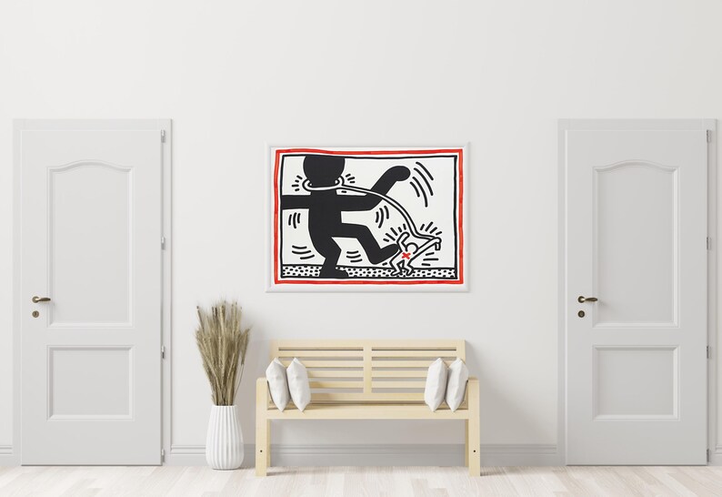 May include: A framed artwork featuring a black and white graphic of a figure with a leash, walking another figure. The artwork has a red border and is displayed above a wooden bench with white pillows. The room has white doors and walls.