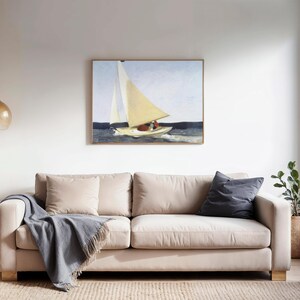 Op de afbeelding: Een beige bank met twee vierkante kussens en een donkergrijze deken. Een ingelijst schilderij van een zeilboot hangt boven de bank. Een kleine bijzettafel en een hanglamp zijn ook zichtbaar.