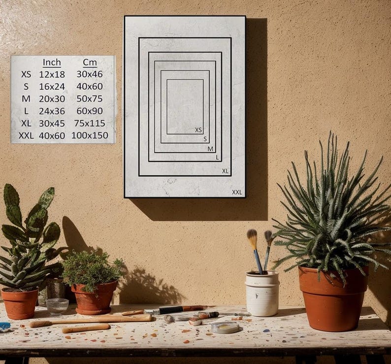 May include: A framed artwork with a series of nested rectangular borders, labeled with sizes XS to XXL. A chart displays inch and centimeter measurements for each size. The artwork is displayed on a beige wall.