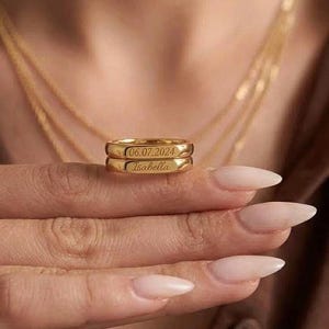 Peut inclure: Deux bagues dorées superposées. La bague supérieure porte la date "06.07.2024" gravée, et la bague inférieure porte le nom "Isabella". Les bagues sont sur le doigt d'une personne, avec un collier doré en arrière-plan.
