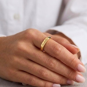 May include: A gold-colored ring with the name "Isabella" engraved on the band. The ring is worn on a finger, showcasing its polished finish and elegant design. The ring is a simple band style.