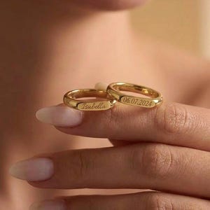 Op de afbeelding: Twee gouden trouwringen. Eén ring is gegraveerd met de naam "Isabella", de andere met de datum "06.07.2024". De ringen worden op de vinger van een persoon getoond.