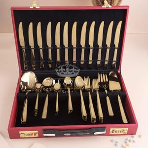 May include: A gold-colored flatware set, including knives, forks, and spoons, is neatly arranged inside a red case with a black interior. The case has a gold clasp and a combination lock. The brand name "HEAVAN Queen" is visible.