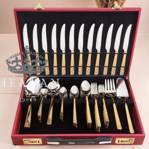 Könnte beinhalten: Ein roter Besteckkoffer mit schwarzem Innenraum, der ein komplettes Set aus gold- und silberfarbenem Besteck enthält. Das Set umfasst Messer, Löffel, Gabeln und Servierutensilien. Der Koffer ist mit einem goldenen Verschluss verschlossen und trägt den Text "HEAN Queen".