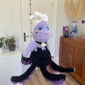 Può includere: Bambola all'uncinetto fatta a mano di una strega del mare, con corpo viola, capelli bianchi e un vestito nero con tentacoli viola. La bambola ha occhi blu e un fiore giallo. Un giocattolo di peluche unico e stravagante.