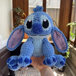 May include: A handmade blue crochet plush toy with large ears, black eyes, and a tuft of hair. The plush toy is a character from a popular animated film. The toy is held in a person's hand.