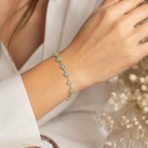 以下が含まれることがあります： 小さな水色の花形のチャームが連なったゴールドのチェーンブレスレット。それぞれの花には小さな黄色の中心部があり、繊細なゴールドのリンクチェーンで留められており、手首の周りに一貫した花柄のデザインを作り出しています。