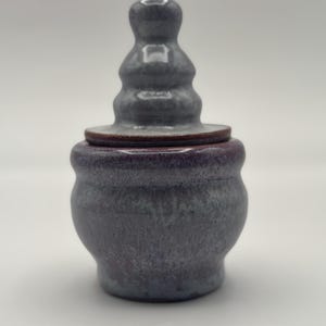 May include: A small, gray ceramic jar with a lid shaped like a spire. The jar has a purple glaze and a textured surface.