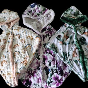 May include: Three hooded capes with textured fabric. The left cape features a rainbow pattern with tan trim. The centre cape displays a purple floral print with white trim. The right cape shows a forest animal scene with green trim.