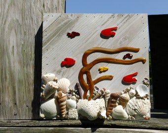 May include: A wooden wall art piece featuring a seascape scene with red and yellow glass fish, brown glass seaweed, and a variety of seashells.