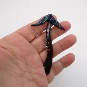 May include: A black hair clip with a decorative iridescent top. The clip is designed to hold hair in place.