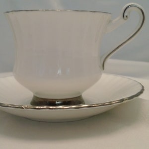 Puede incluir: Una taza de té y un platillo blancos con un borde plateado. La taza tiene un asa delicada y el platillo tiene un borde festoneado.